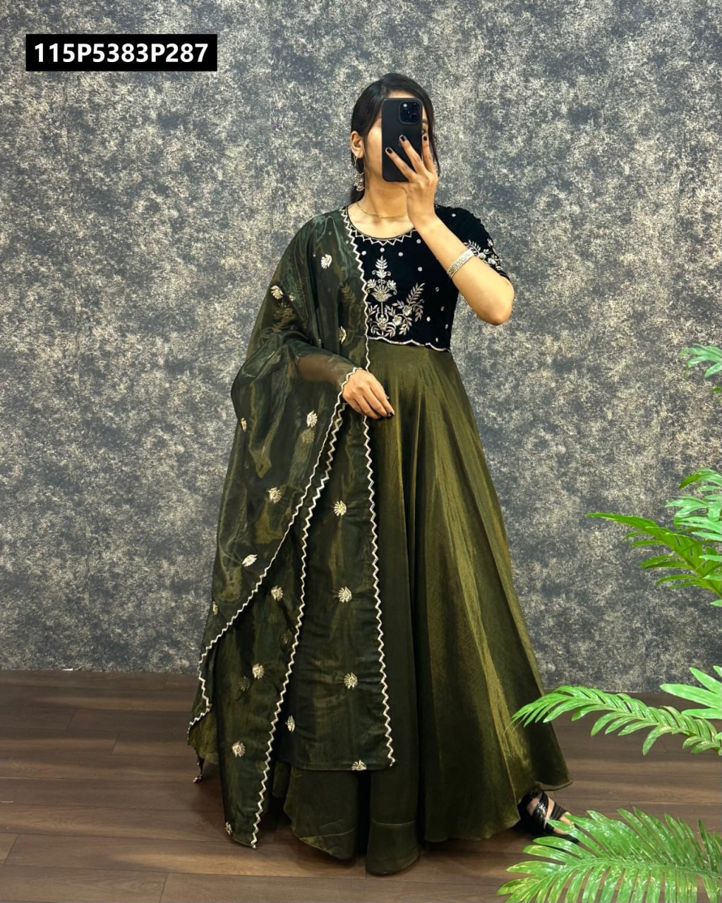Full body shot of a woman modeling an olive green embroidered velvet gown paired with a matching tissue net dupatta, against a textured grey wall.