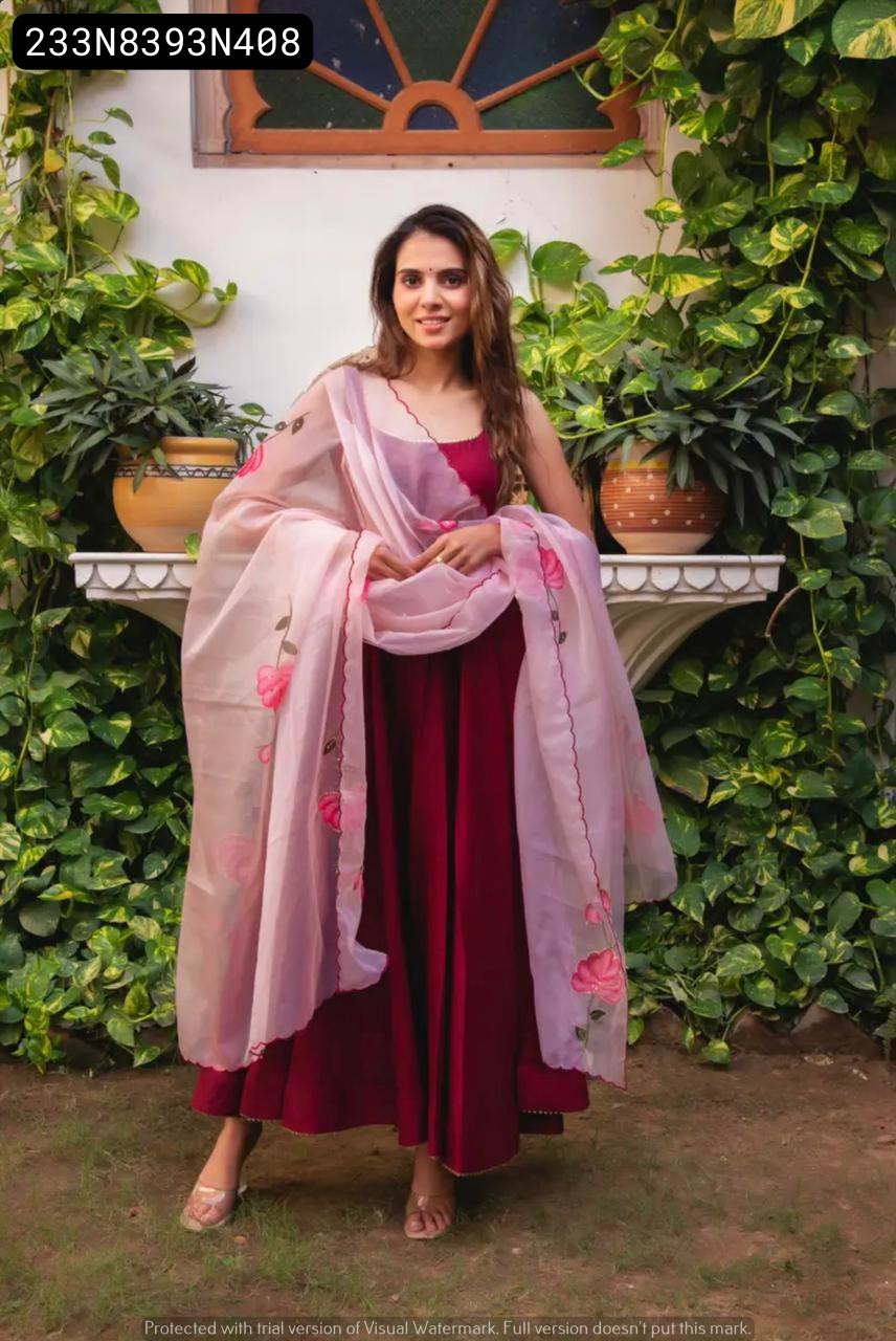 Model in a deep maroon Chinon Anarkali suit with a light pink pure organza digital printed dupatta, posing gracefully against a lush green leafy background.