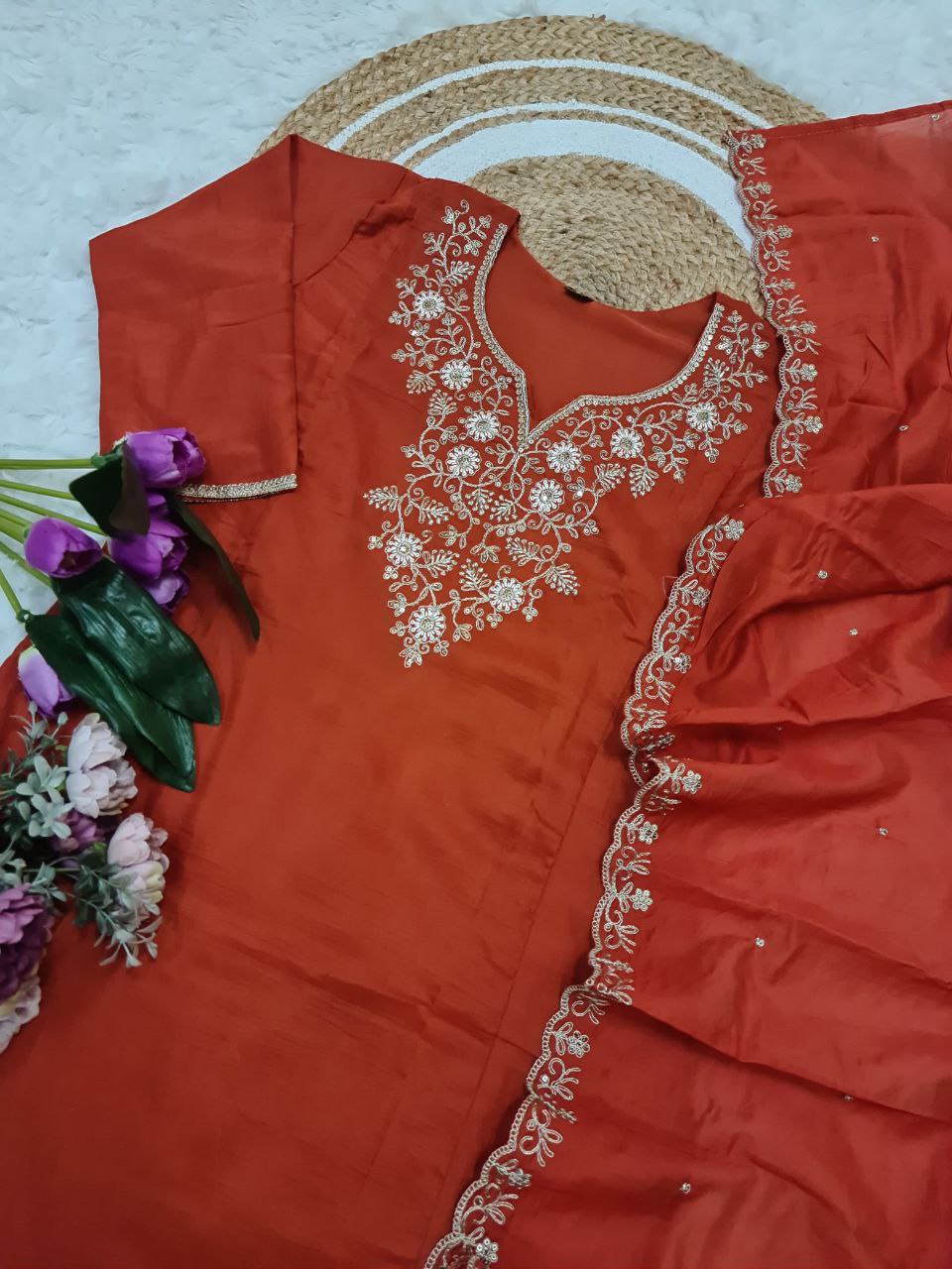 Flat lay of the orange embroidered Viscose Chinon kurta, pant, and Chanderi Cotton dupatta, laid out on a textured mat with purple flowers.