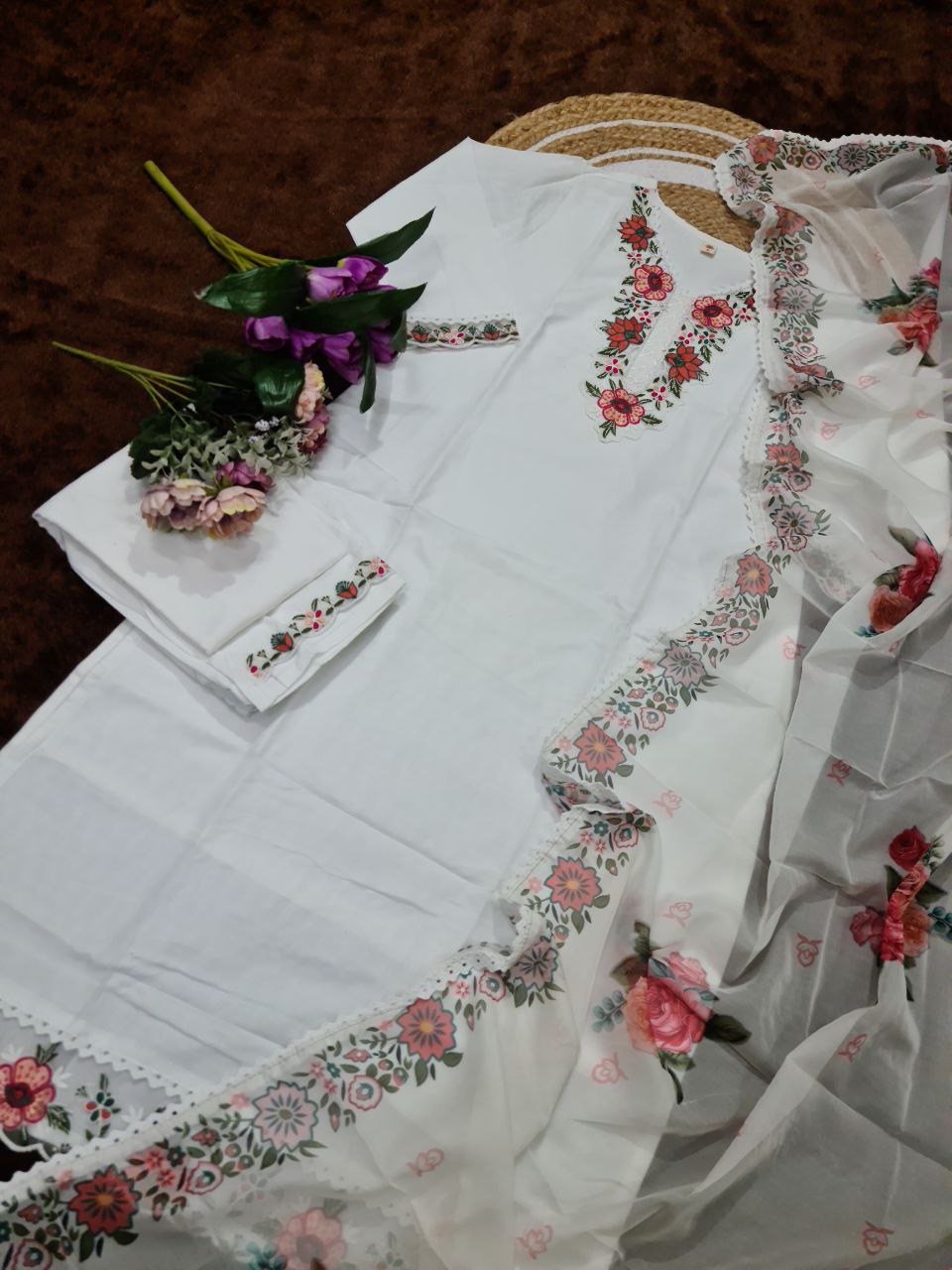 Overhead flat lay of the TRENDBUY white embroidered kurti set, including the kurti, pant, and dupatta, styled with flowers on a rustic mat.