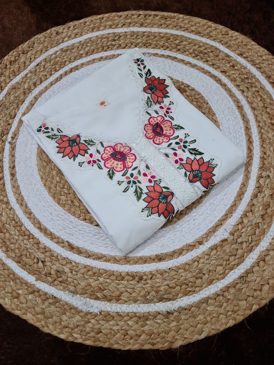 Flat lay of the neatly folded TRENDBUY white embroidered kurti, focusing on the intricate floral embroidery on the neckline, on a textured mat.