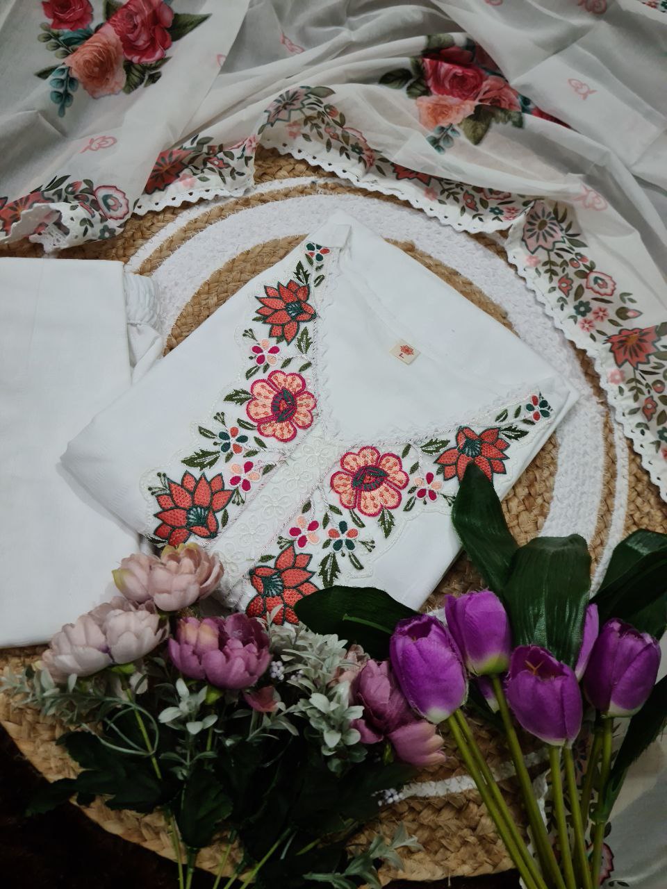 Overhead flat lay of the white embroidered kurti, pant, and taby dupatta ensemble, adorned with vibrant fresh flowers for aesthetic appeal.