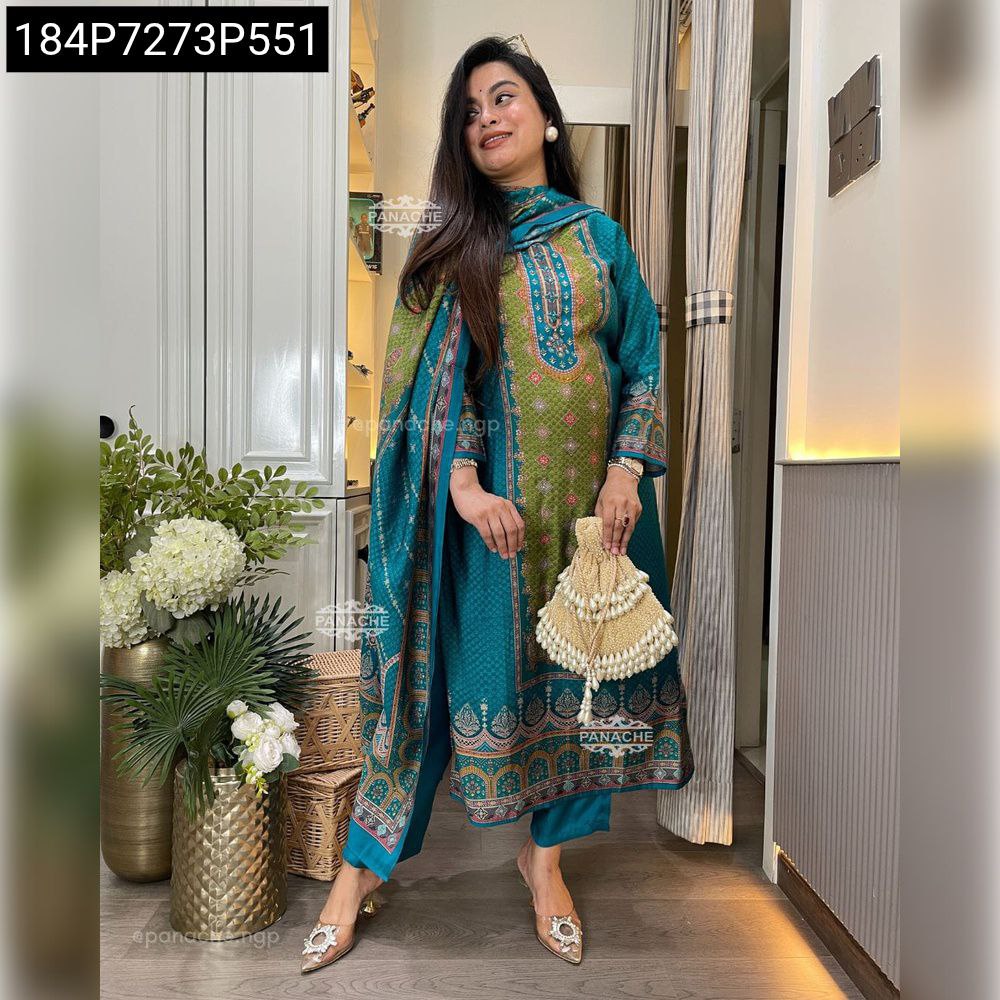 Full body shot of a woman in a teal and green KitKat silk suit set with intricate handwork, holding a beaded potli bag, in a modern living space.