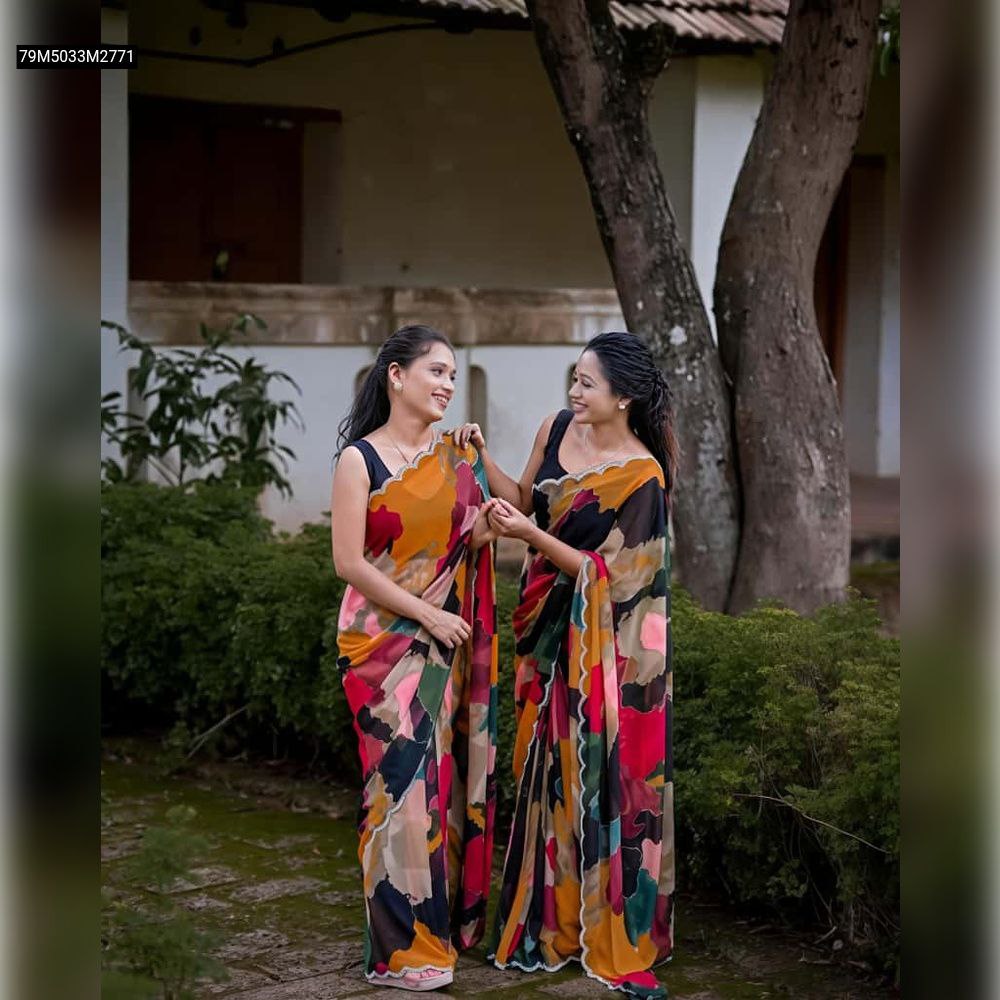 Two models in full-length mosaic print georgette sarees with scalloped borders, one with a yellow/orange base and one with a black base, standing in a garden setting, smiling and interacting.