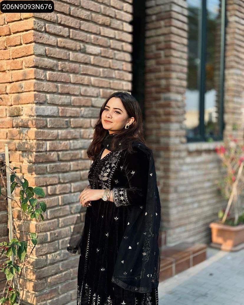 Woman in black embroidered velvet Anarkali gown, smiling, against a brick wall background.