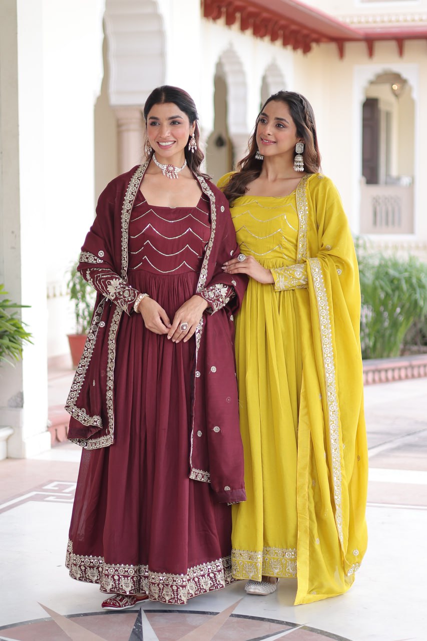 Two women, one in maroon anarkali gown with dupatta, one in yellow, posing gracefully outdoors.
