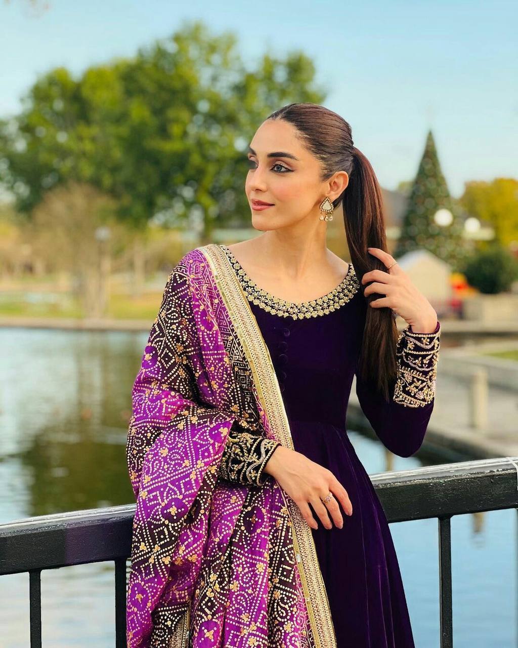 Elegant woman in a deep purple velvet gown with ornate dupatta, posing outdoors by a lake.
