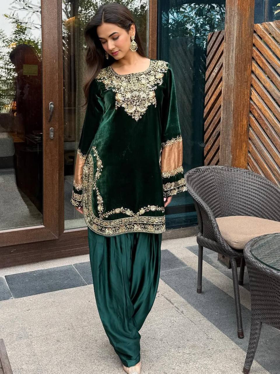 Full body shot of a woman modeling a dark green velvet kurta salwar suit with gold sequence embroidery, standing outdoors.