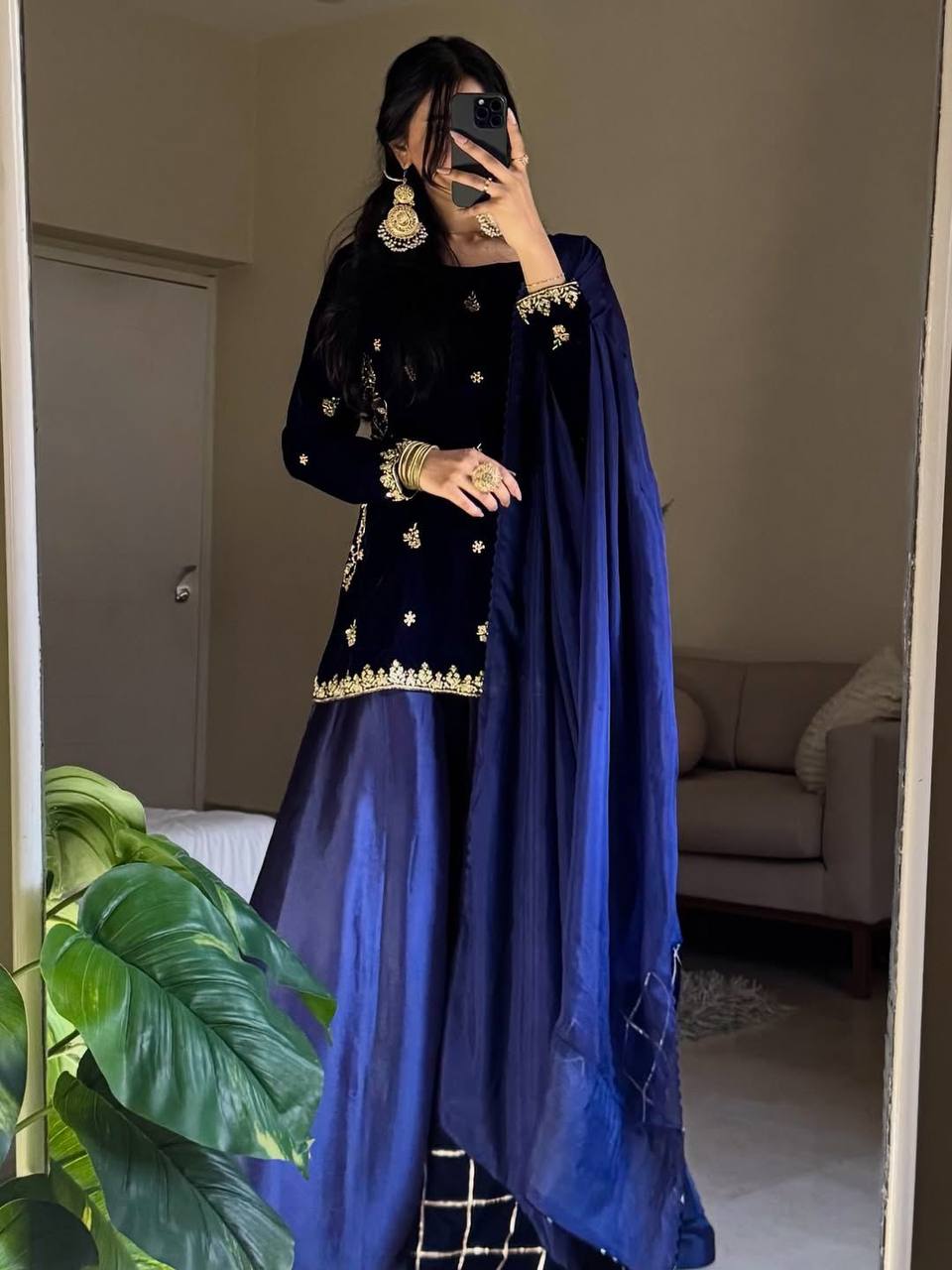 Full shot of a woman in a blue velvet embroidered plazzo suit, holding a phone, in a mirror.