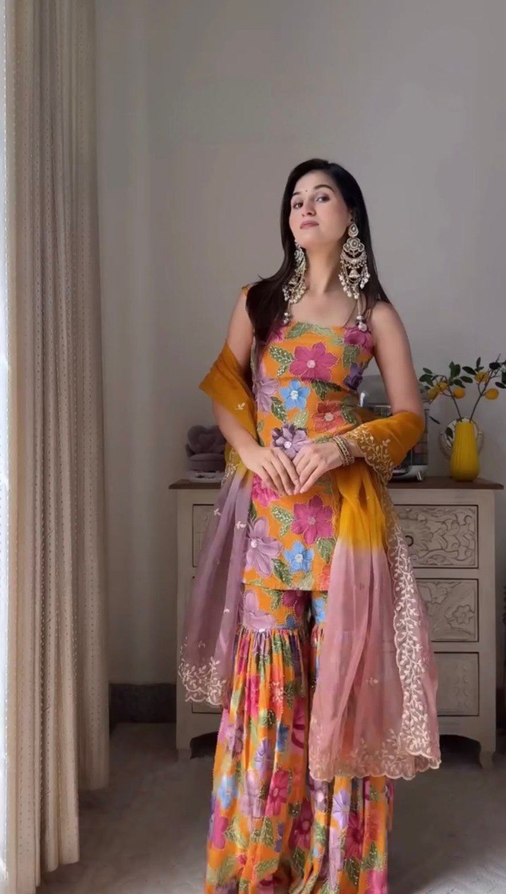 Woman wearing a vibrant orange floral print sleeveless top and flared sharara pants with a yellow and pink ombre dupatta, accessorized with statement earrings.