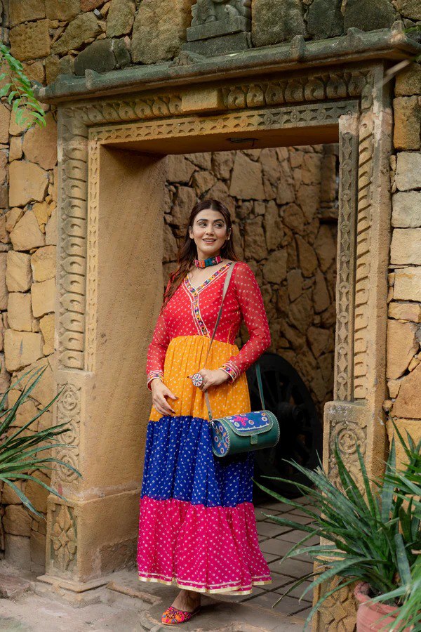 Woman posing in a vibrant, multi-colored georgette ruffle gown with mirror work and embroidered border, standing in an ancient doorway.