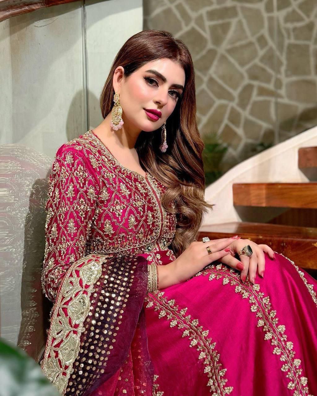 Model wearing a vibrant pink Anarkali gown with heavy gold embroidery and sequins, sitting on stairs.