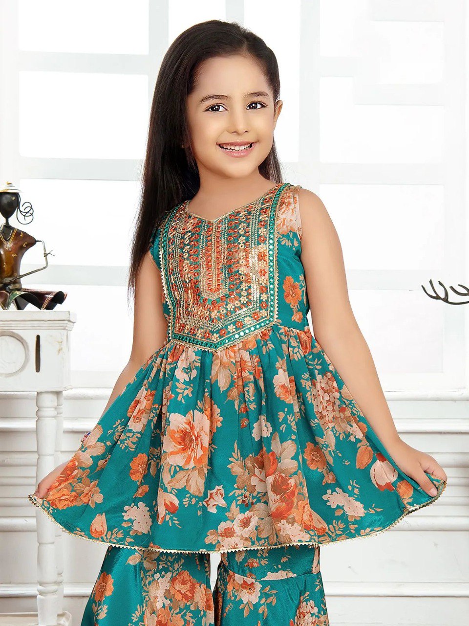Young girl smiling, wearing a teal floral print sleeveless top with intricate embroidery and sequins at the neckline, paired with matching sharara pants.