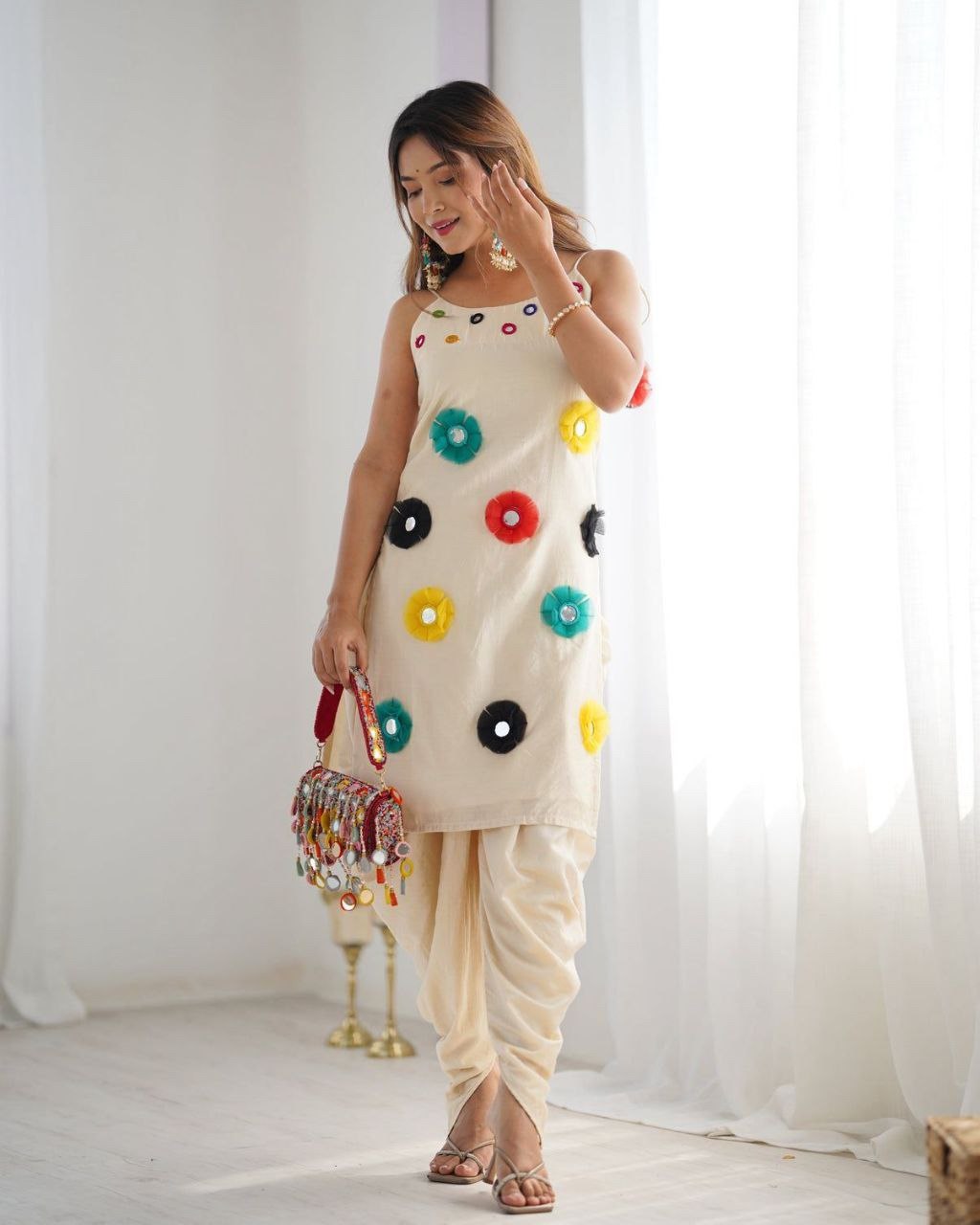 Woman wearing a beige Navratri special dhoti kurta set with colorful mirror work, holding a matching embellished bag, looking down.