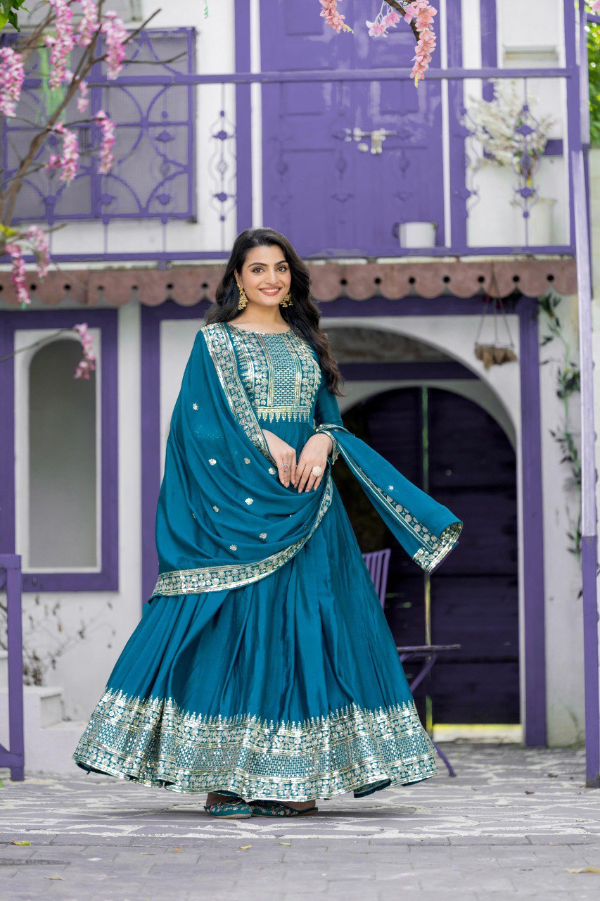 Full shot of woman in elegant teal Chinon silk Anarkali gown with 5mm sequence embroidery, smiling against a purple building.