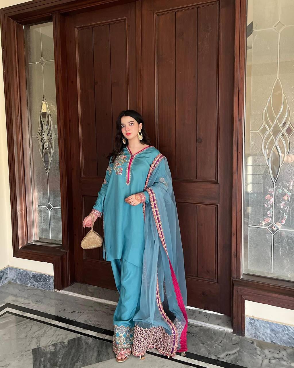TRENDBUY model showcasing a full-length view of the teal embroidered Kurta, Palazzo, and Net Dupatta set, standing elegantly by wooden doors.