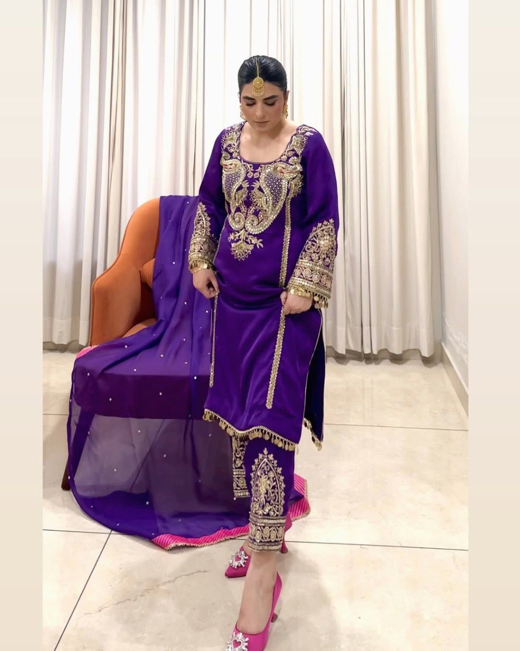 Model showcasing a purple georgette suit set with heavy gold embroidery and sequence work, standing next to an armchair.