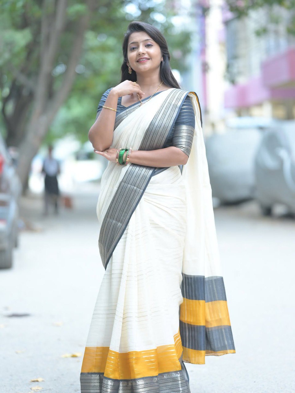 Smiling woman posing gracefully in an off-white Mysore silk saree with black and gold padding border, outdoor setting.