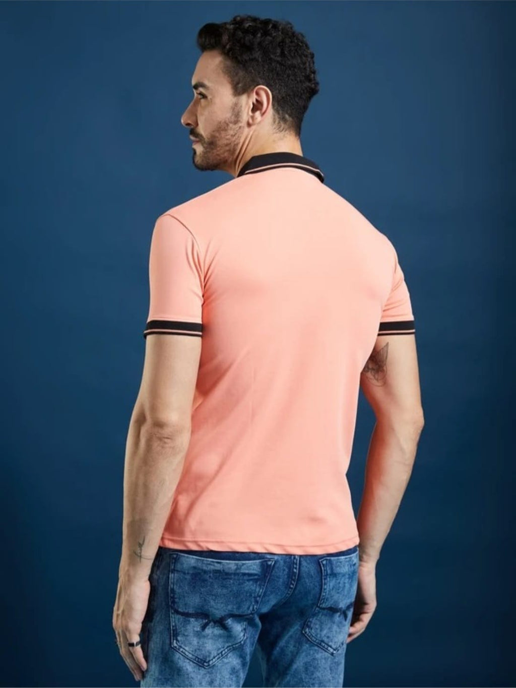 Rear view of a man wearing a peach V-neck polo t-shirt with black collar and cuff trim, paired with blue jeans, against a blue background.