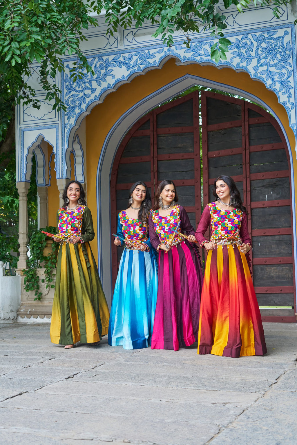Four women in vibrant Kutchi Gamthi embroidered lehenga cholis against a traditional Indian arched doorway, smiling brightly.