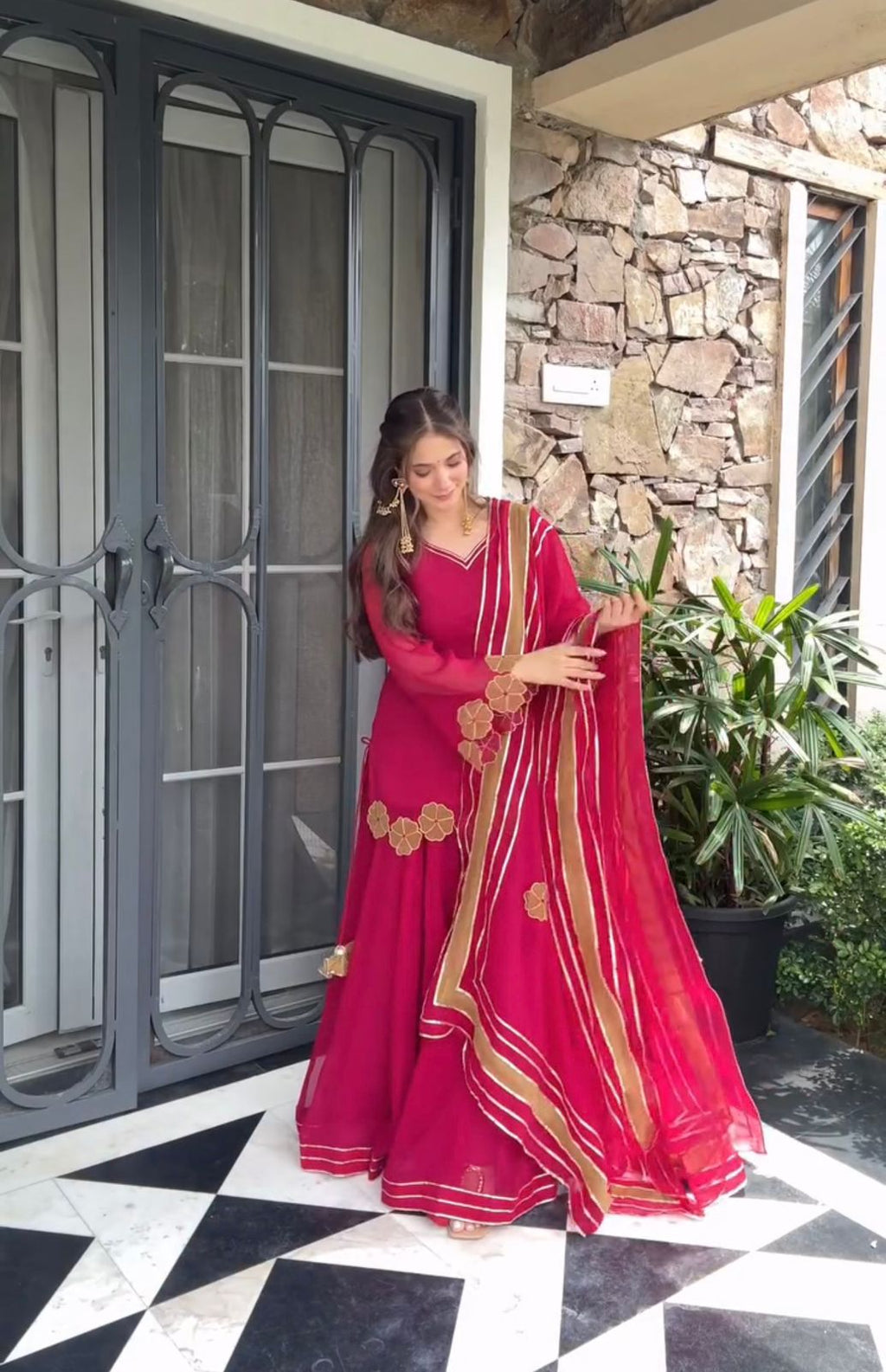 Full shot of a woman in a fuchsia embroidered plazzo set with dupatta, posing elegantly against a stone wall.