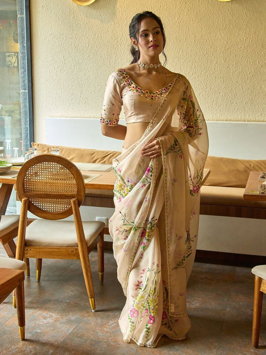 Model in cream Tebby Silk floral handwork saree with embroidered V-neck blouse, posed in a chic cafe setting.