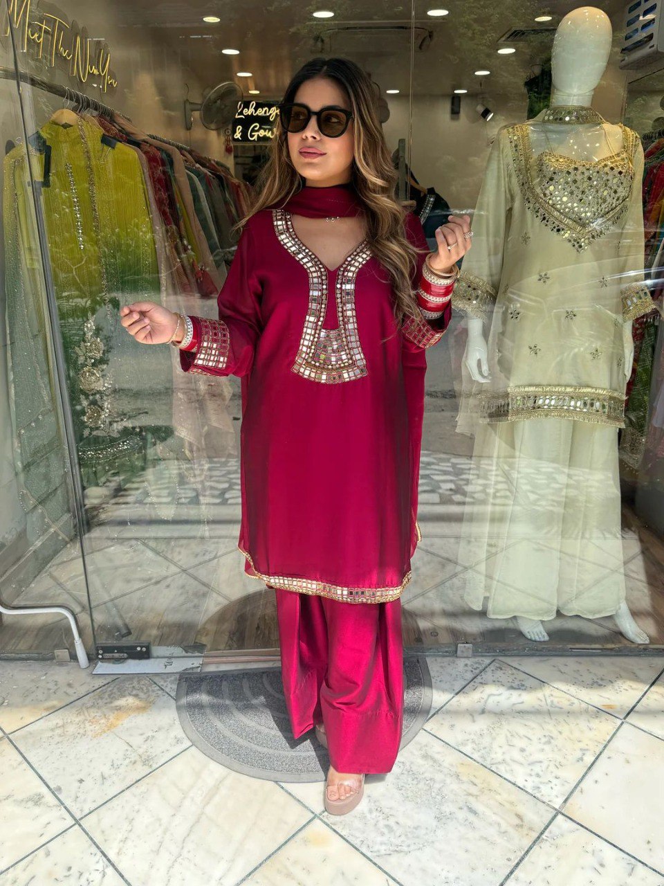 Woman confidently posing in a maroon Rangoli silk kurti pent dupatta set with real mirror work, outside a boutique.
