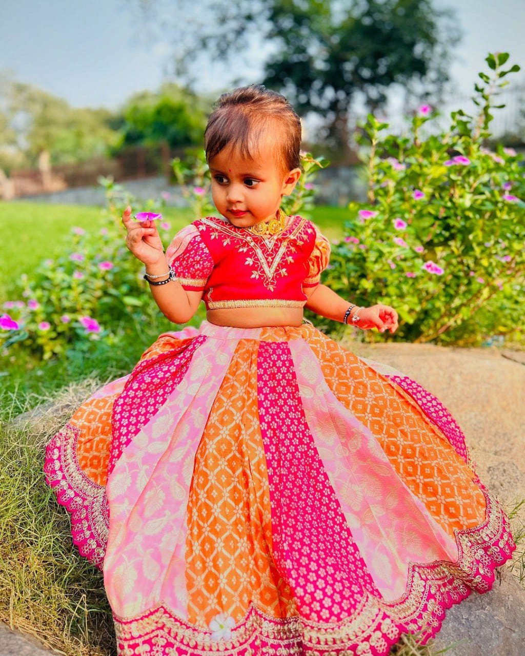 Adorable toddler girl wearing a multi-color handwoven lehenga choli set, sitting outdoors in a garden setting.