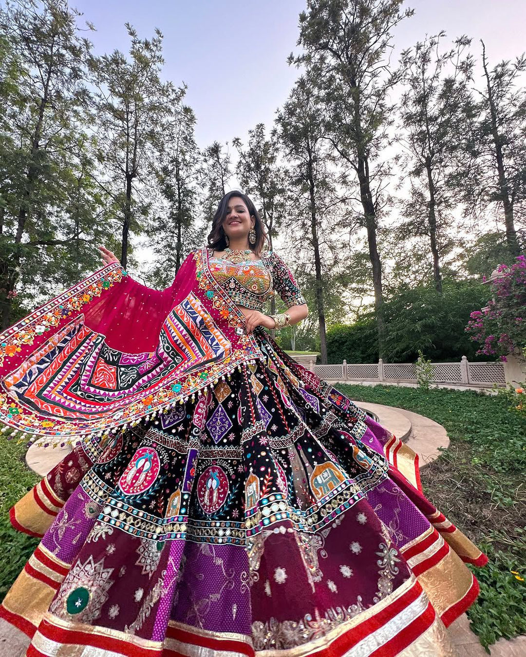 Smiling woman in a vibrant black, purple, and red Navratri Chaniya Choli with real mirror work, twirling gracefully in a garden setting.