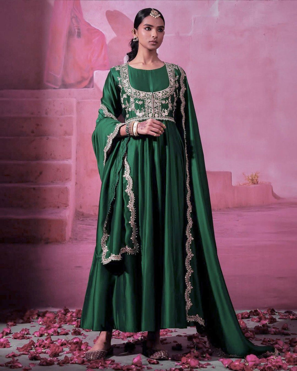 Full shot of a model elegantly posing in a stunning green chinon silk gown with an embroidered koti and matching dupatta, against a pink background with rose petals.