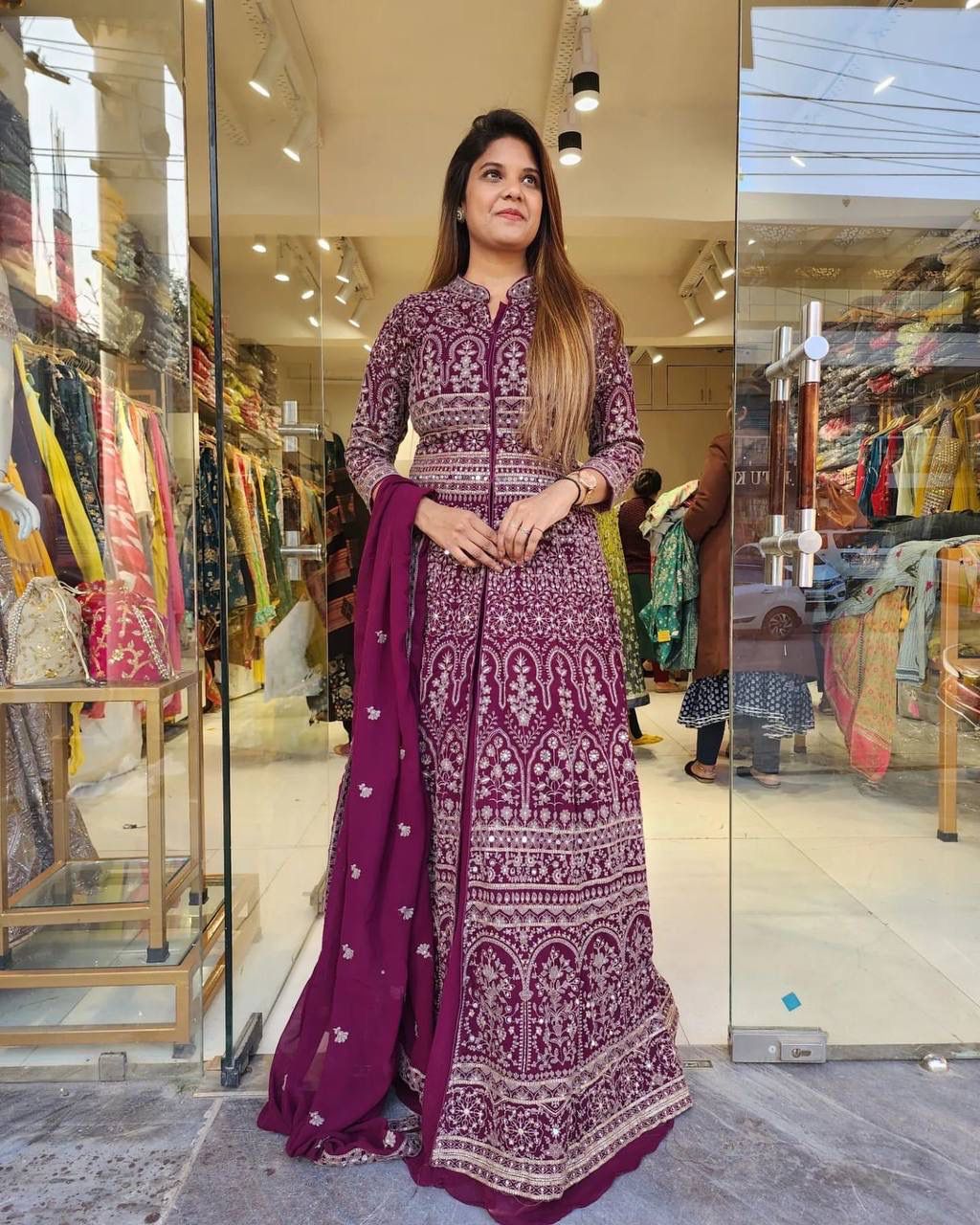 Woman in deep purple designer georgette koti lehenga set with intricate gold and mirror embroidery, standing elegantly.