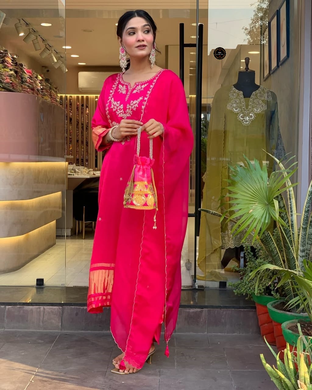 Full shot of a model wearing a hot pink viscose silk kurti set with exquisite embroidery, holding a matching embroidered potli bag, standing in front of a modern store.
