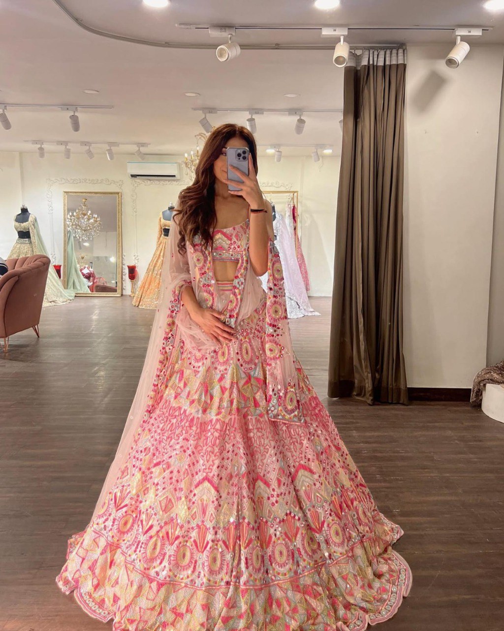 Model in pink multi-color embroidered lehenga choli with mirror work, taking a mirror selfie in a boutique.