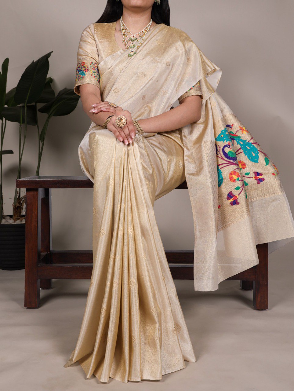 Full view of woman elegantly draped in a cream Tissue Paithani Saree with intricate Zari weaving and a colorful peacock motif on the pallu, sitting gracefully.