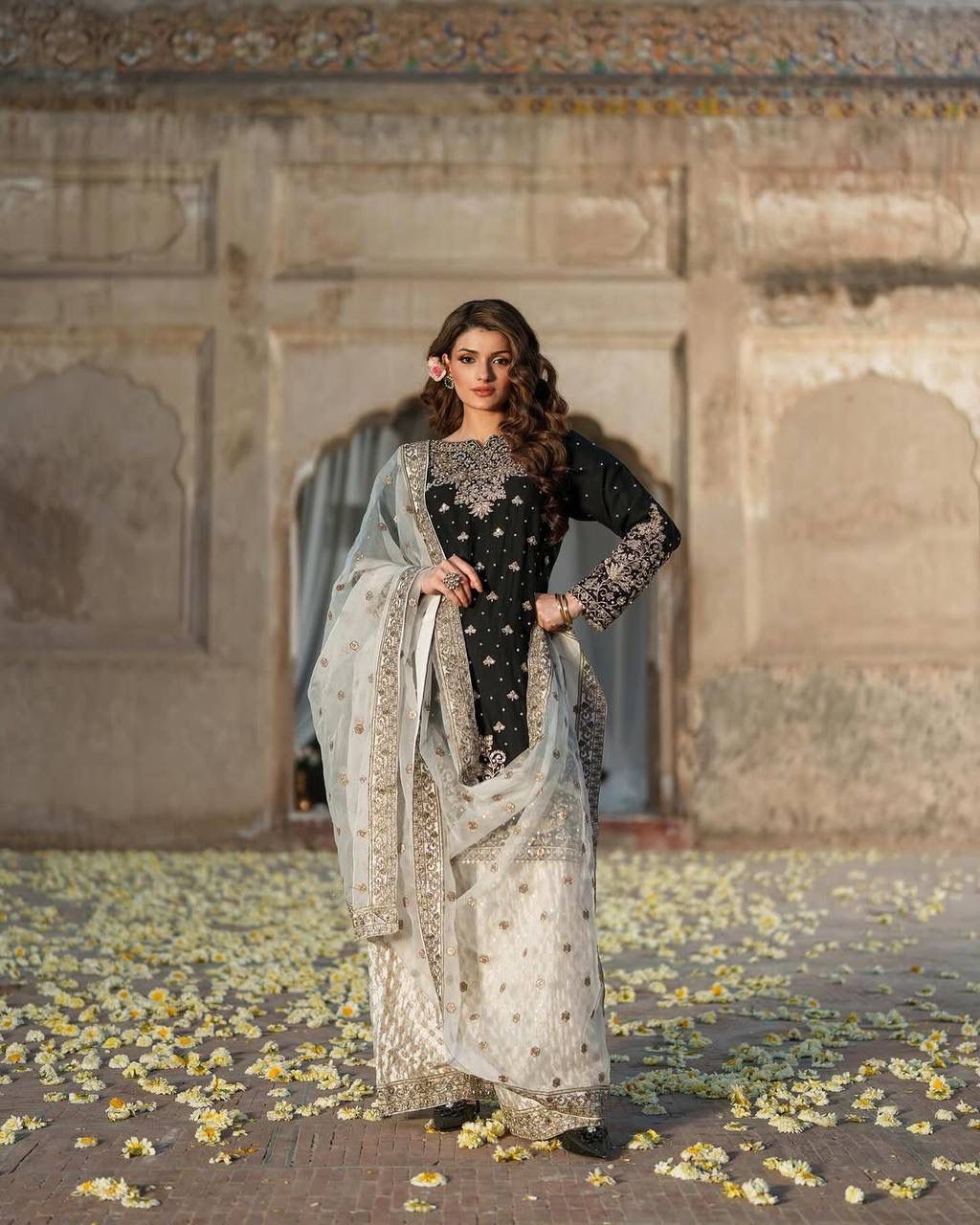 Model showcasing a black embroidered Chinon silk plazzo suit set with white dupatta, standing gracefully in an archway with scattered petals.