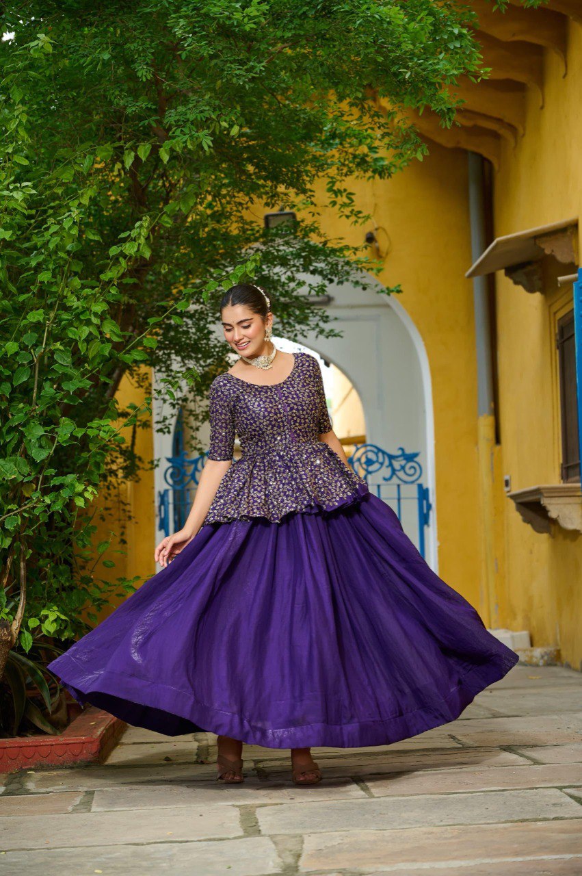 Elegant model in purple sequined peplum blouse and flowing purple lehenga, striking a pose outdoors with a joyful expression.