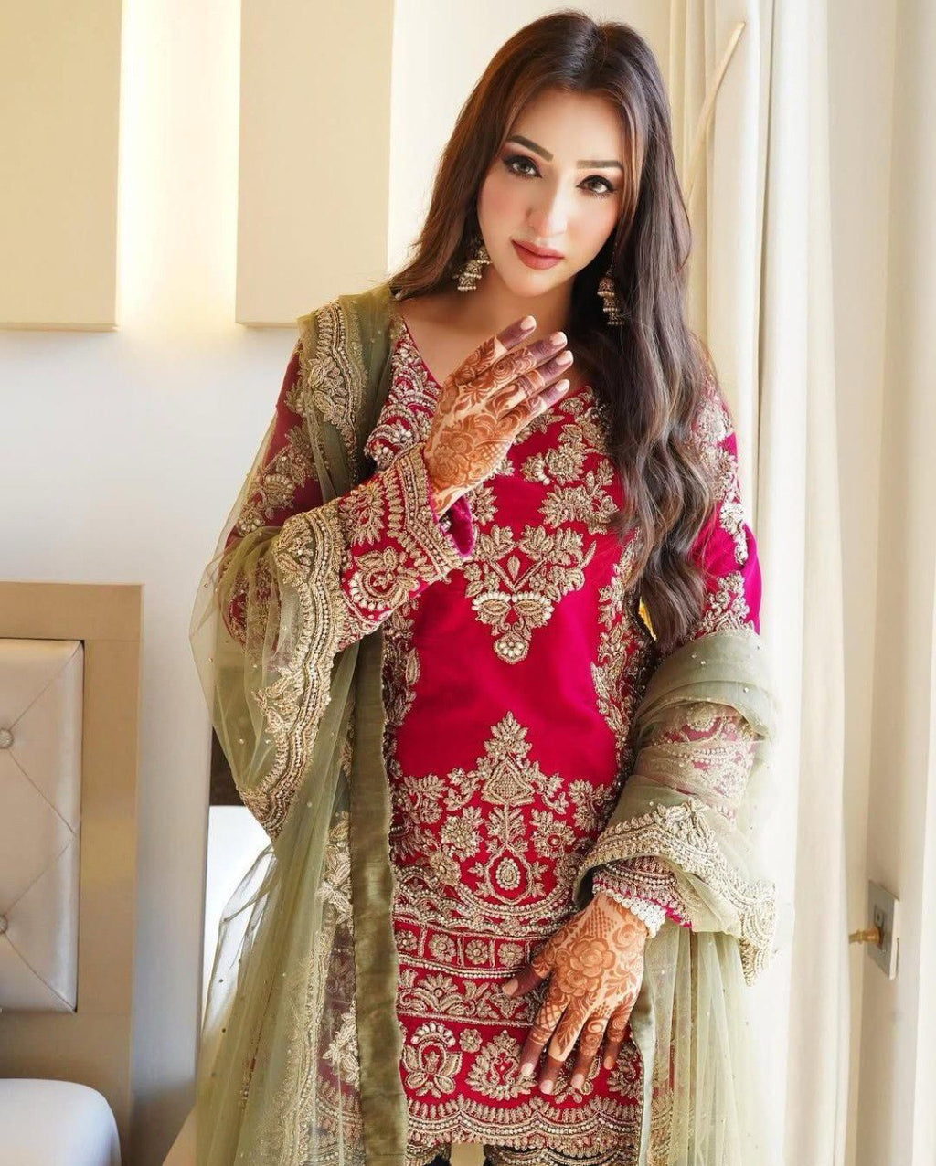 Full-length shot of a woman wearing a red embroidered sharara top with an olive green net dupatta, showcasing intricate mehndi designs on her hands.