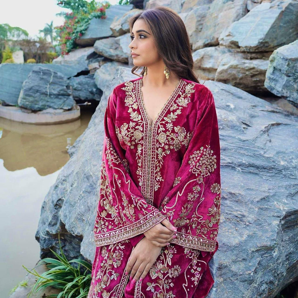 Elegant woman in magenta velvet kurti plazzo set with gold sequin embroidery, posing against rocks.