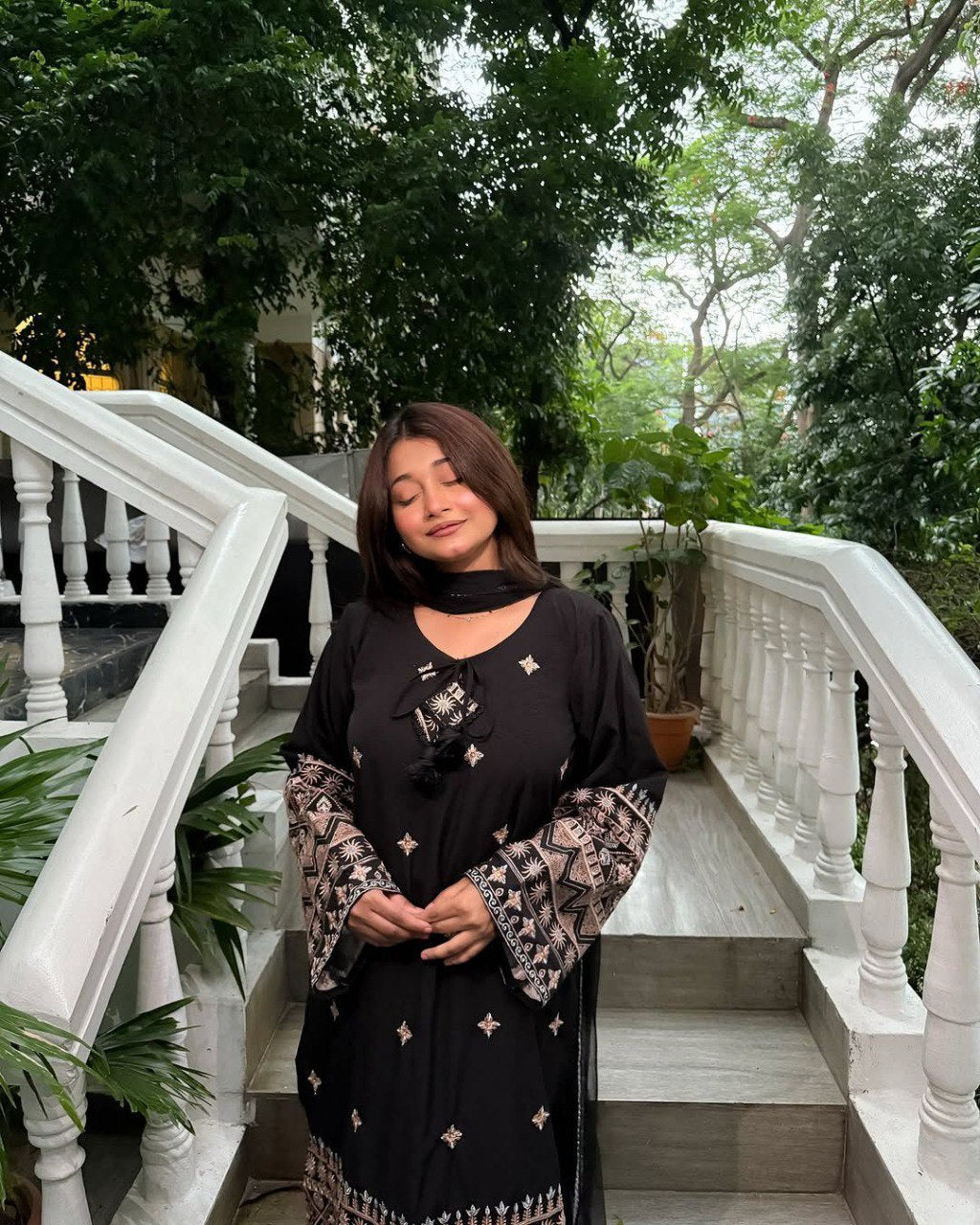 Model in black Roman Silk Cotton kurti plazzo set, standing on stairs, elegant pose with subtle smile.