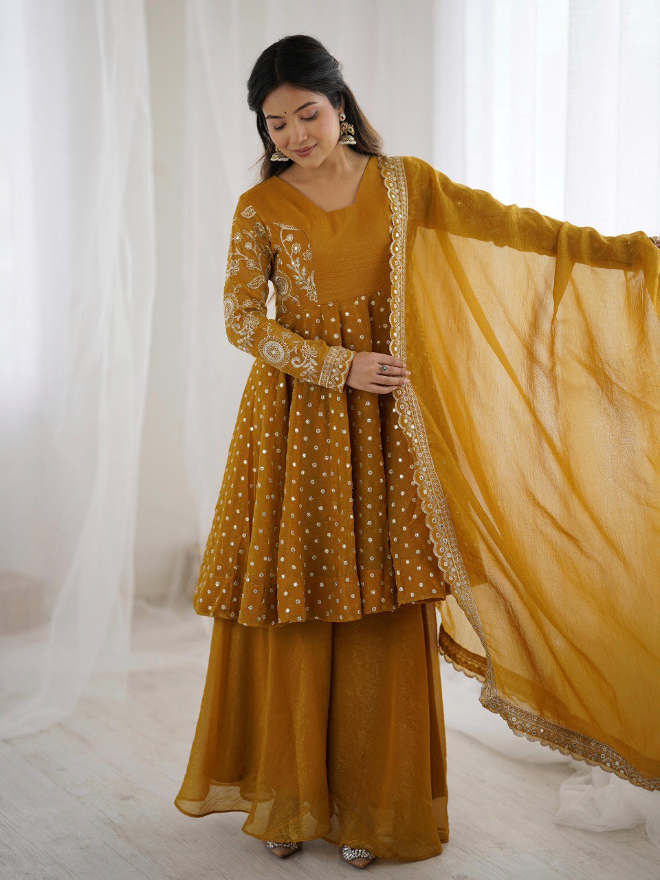 TRENDBUY woman gracefully posing in a mustard yellow crunchy silk sharara suit with embroidery.