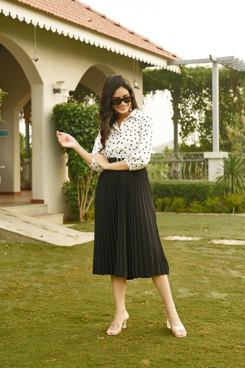 Woman in black heart print top and black pleated midi skirt, smiling outdoors.