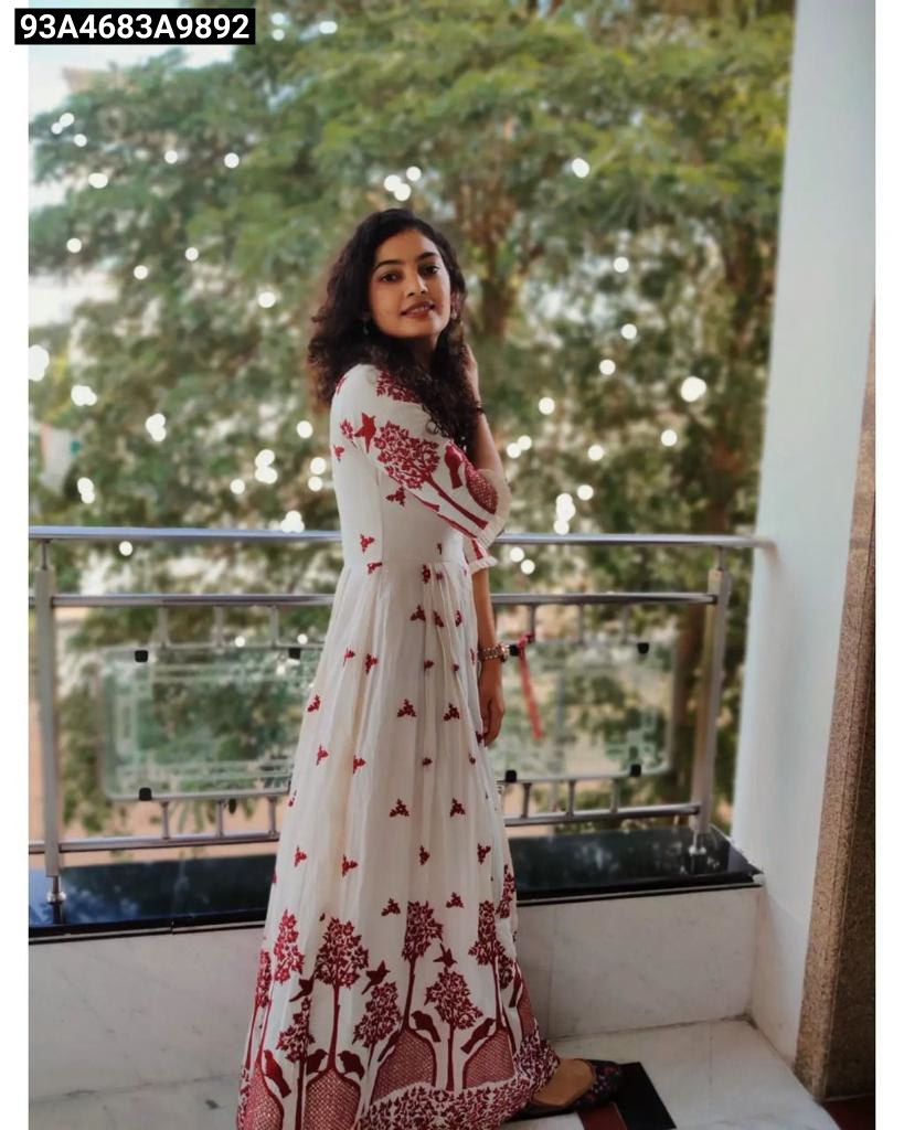 Woman in a white dress with red patterns standing on a balcony with greenery in the background