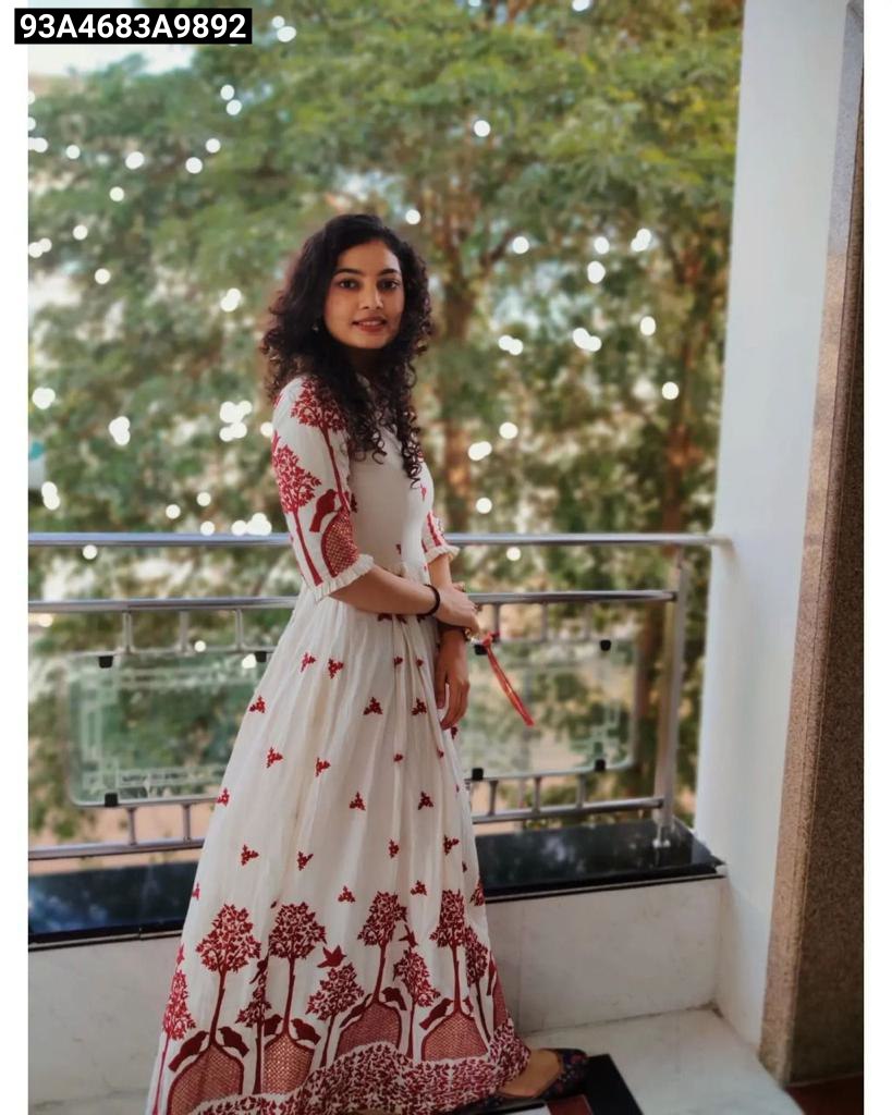 Woman in a white dress with red patterns standing on a balcony with greenery in the background