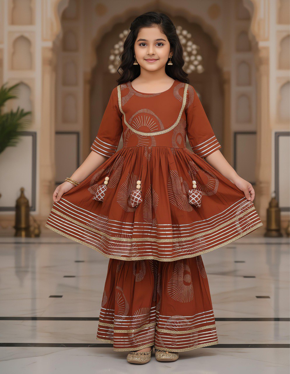 Young girl in a rust brown designer kurta kediya and sharara set, smiling, holding out skirt in a traditional palace setting.