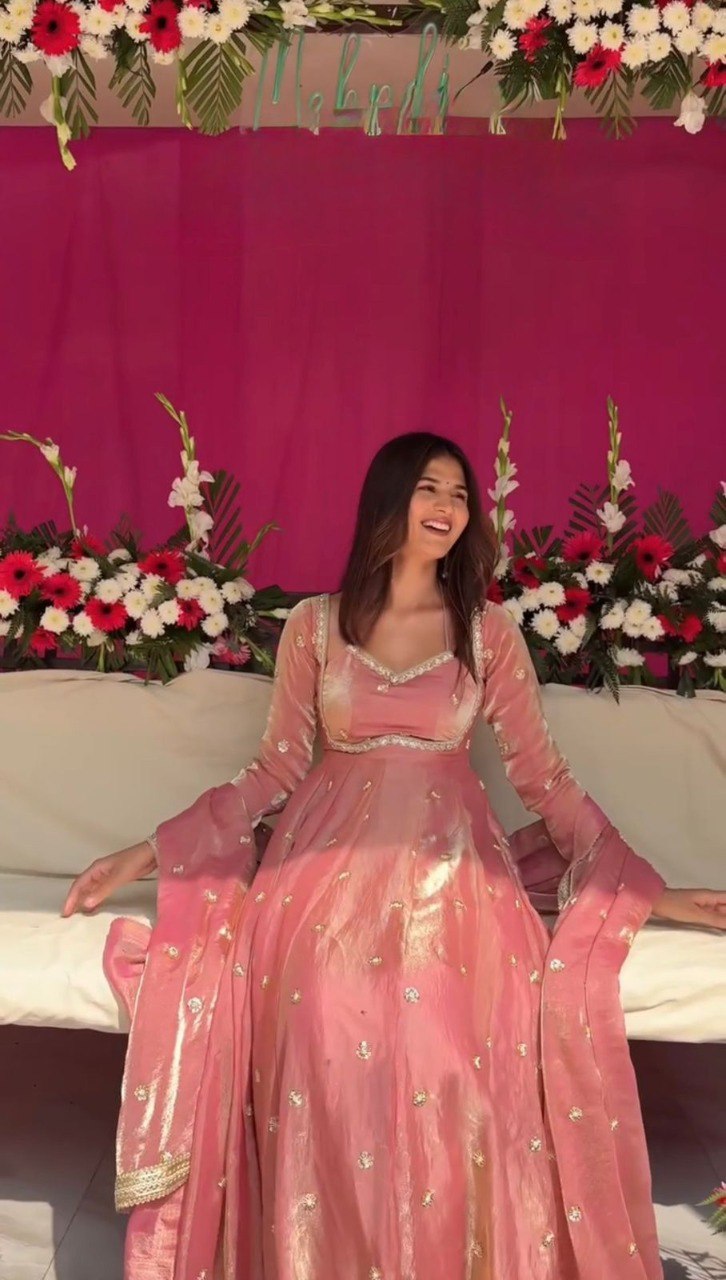 Smiling model in an elegant blush pink Fendy Silk Anarkali suit set with sequin work, seated on a white couch.