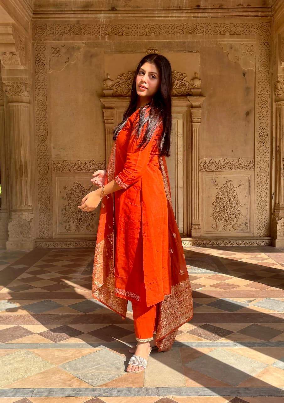Model wearing an elegant Orange Roman Silk suit set with embroidered neckline and Chanderi dupatta, posing outdoors in a heritage location.