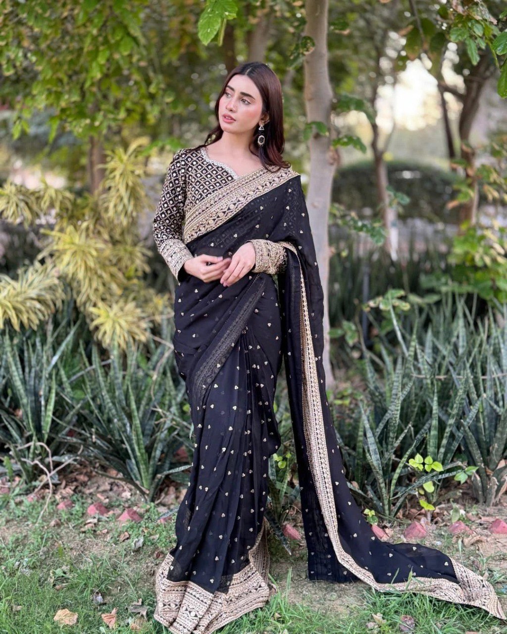 Full shot of a model gracefully posing in a black Tibby silk saree with golden sequin and thread work in a lush garden.