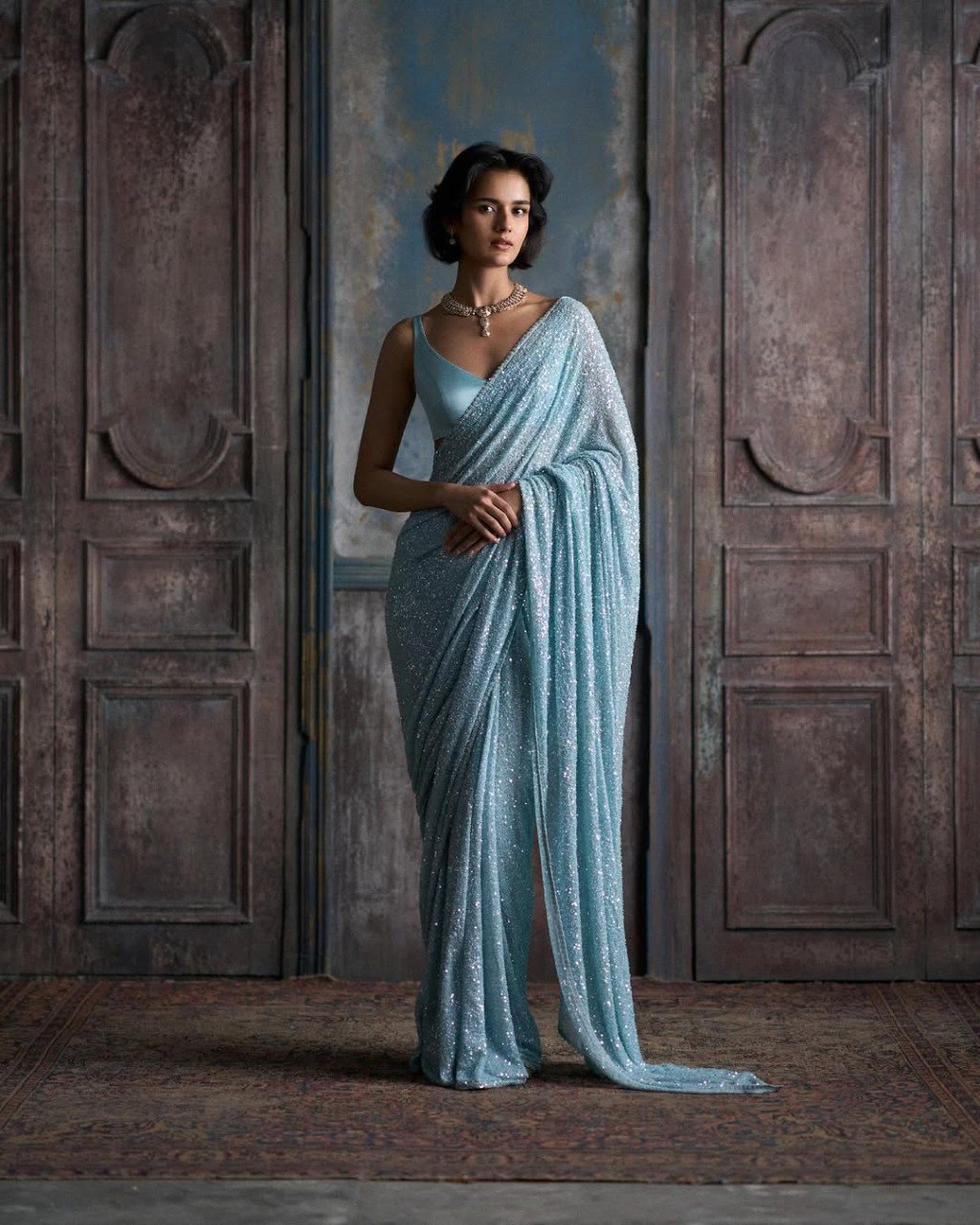 Model wearing a stunning light blue faux georgette sequin saree with a matching plain silk blouse and necklace, posing against an antique wooden door.
