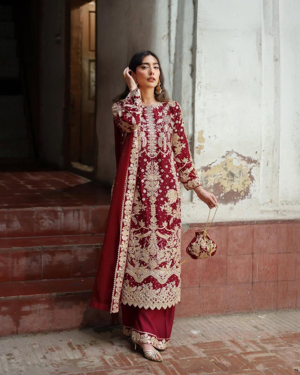 Model gracefully posing in a deep red embroidered sequence designer suit with a matching potli bag.
