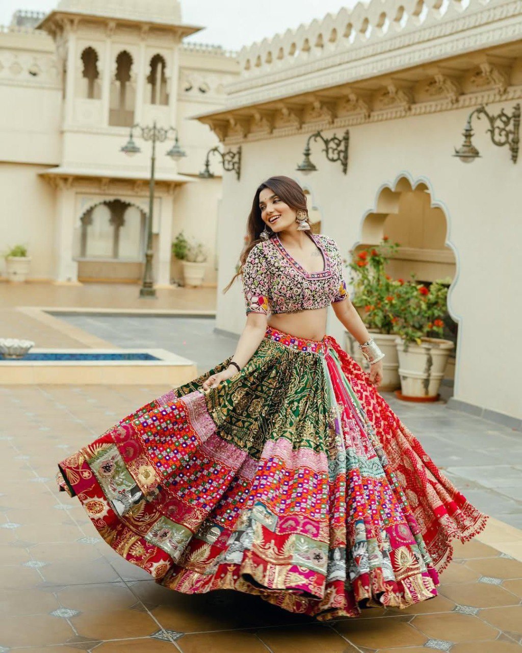 Smiling woman spinning joyfully in a vibrant multicolor Navratri Chaniya Choli with real mirror work, set against a traditional Indian palace backdrop.